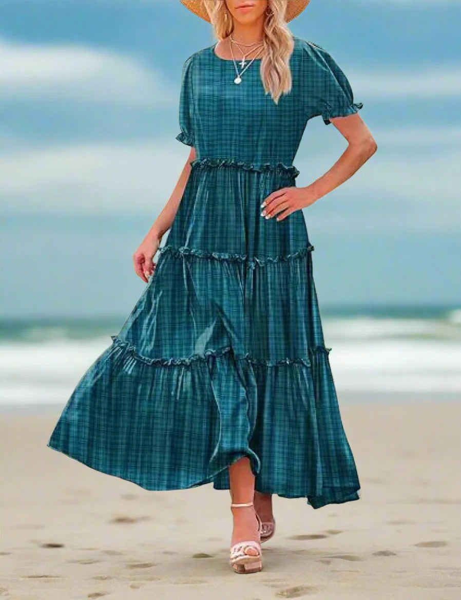 Woman wearing a teal plaid dress standing outdoors with a stone building and plants in the background.