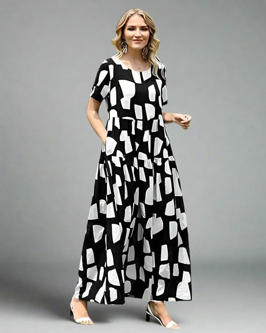 Woman wearing a black and white patterned dress standing outdoors on grass with trees and a white fence in the background.