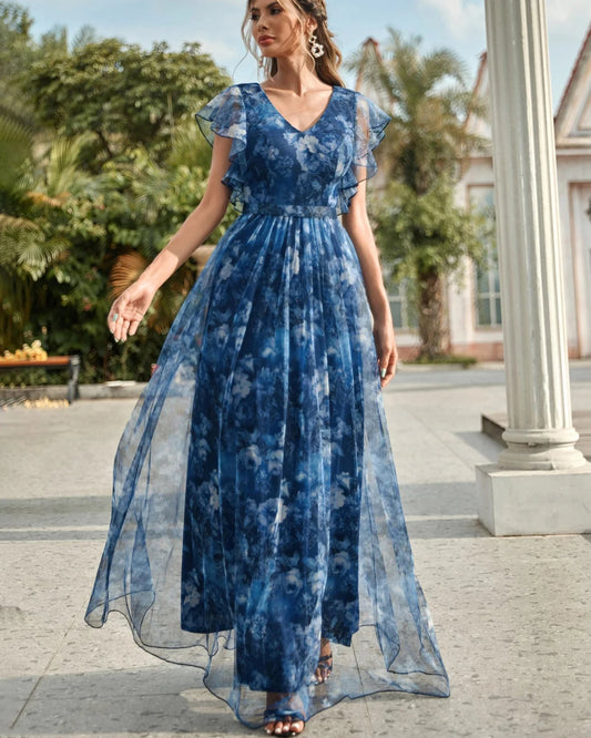 Woman wearing a blue floral dress standing outdoors with greenery and a building in the background